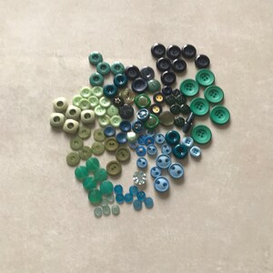 May include: A collection of green, blue, and black buttons in various sizes and shapes. The buttons are arranged in a circular pattern.
