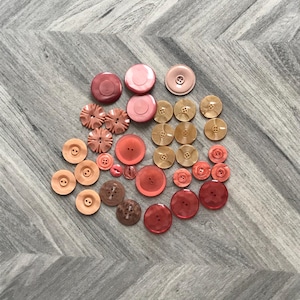 May include: A collection of various sizes and shades of pink and red buttons. The buttons include round, flower-shaped, and flat designs. Some buttons have two or four holes, while others have a shank. The buttons are arranged on a gray and white patterned surface.