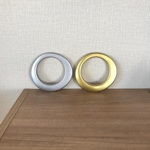 May include: Two curtain grommets, one silver and one gold, are displayed on a wooden surface against a textured, off-white wall. The grommets are circular with a smooth, metallic finish, ideal for drapery.