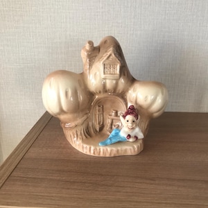 May include: A ceramic ornament depicting a fantasy house with a small figure. The house is beige with a small window and door. The figure wears a red hat and blue scarf. The ornament sits on a wooden surface.