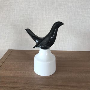 May include: A black bird ornament with a glossy finish, mounted on a white, cylindrical base. The bird has a streamlined, contemporary design. The sculpture is displayed on a wooden surface.