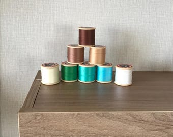 May include: A collection of colorful spools of thread arranged on a wooden surface. The spools are in various colors, including brown, green, teal, and blue. The thread spools are stacked in a pyramid shape, with a neutral-colored wall in the background.