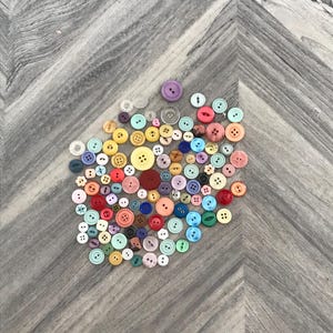 May include: A collection of colorful buttons in various sizes and shades, including red, yellow, blue, and pink. The buttons are scattered on a gray, chevron-patterned surface, creating a visually appealing arrangement for crafts or sewing projects.