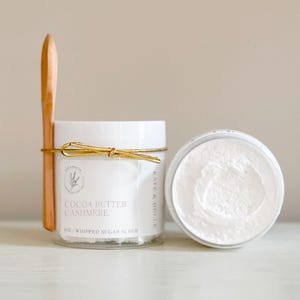 May include: A white jar with a gold lid and a wooden spoon. The jar is labeled "Cocoa Butter Cashmere Whipped Sugar Scrub".