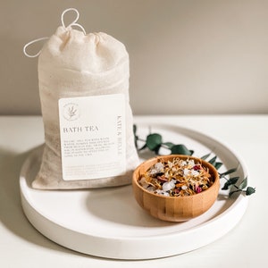 May include: A white muslin bag filled with bath tea, labelled "Bath Tea" and "Kate & Belle". The bag is sitting on a white tray with a small wooden bowl filled with dried flowers, herbs, and salts.