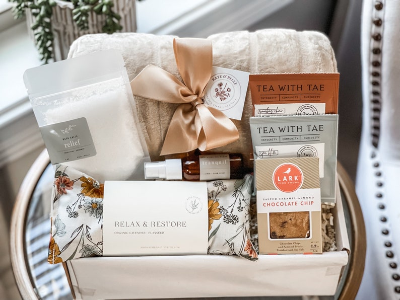 May include: A gift hamper containing a beige plush blanket tied with a satin ribbon, a pouch of bath salts labelled Relief, a small amber spray bottle, two Tea With Tae packets, and a box of Lark Salted Caramel Almond Chocolate Chip cookies.