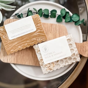 Op de afbeelding: Twee handgemaakte zeepjes op een houten snijplank. De zeep links is een honing- en havermelkzeep met een label waarop staat "Handgemaakte geitenmelkzeep met echte honing, havervlokken en calendula.  Origineel recept.  Handgemaakt en geurend.  www.kateandbelleco.com". De zeep rechts is een witte haver- en honingzeep met een label waarop staat "Haver, melk & honing.  Geitenmelkzeep met havervlokken en honing.  Handgemaakt natuurlijk."