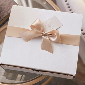 May include: A square white cardboard gift box wrapped with a smooth champagne-coloured satin ribbon tied in a bow. A small rectangular tag with the text Sage & Belle Co. and a floral line drawing is tucked under the ribbon on the top surface.