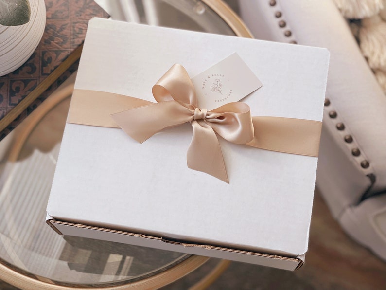 May include: A square white cardboard gift box wrapped with a smooth champagne-coloured satin ribbon tied in a bow. A small rectangular tag with the text Sage & Belle Co. and a floral line drawing is tucked under the ribbon on the top surface.