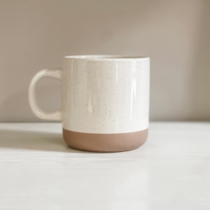Op de afbeelding: Een witte keramische mok met een gespikkeld glazuur en een bruine rand. De mok staat op een wit oppervlak.