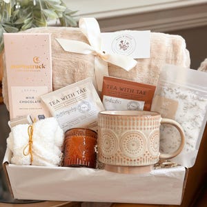 Könnte beinhalten: Eine Geschenkbox mit einer beigen Decke, die mit einem weißen Band gebunden ist, einer rosa Schokoladenstange, Teebeuteln, einer gemusterten Tasse, einer Kerze und einem Beutel Badesalz. Die Box ist weiß und mit weißem Seidenpapier ausgekleidet.