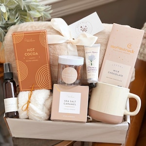 May include: A gift basket containing a tan plush blanket with a white ribbon, a box of salted caramel hot cocoa, a jar of cookies, a tube of French lavender hand cream, a Moonstruck milk chocolate bar, a spray bottle, and a two-tone ceramic mug.