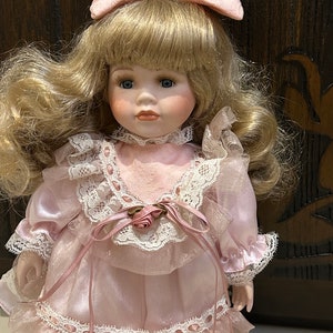 May include: A porcelain doll with blonde hair and a pink dress with lace trim. The doll is wearing a pink bow in her hair.
