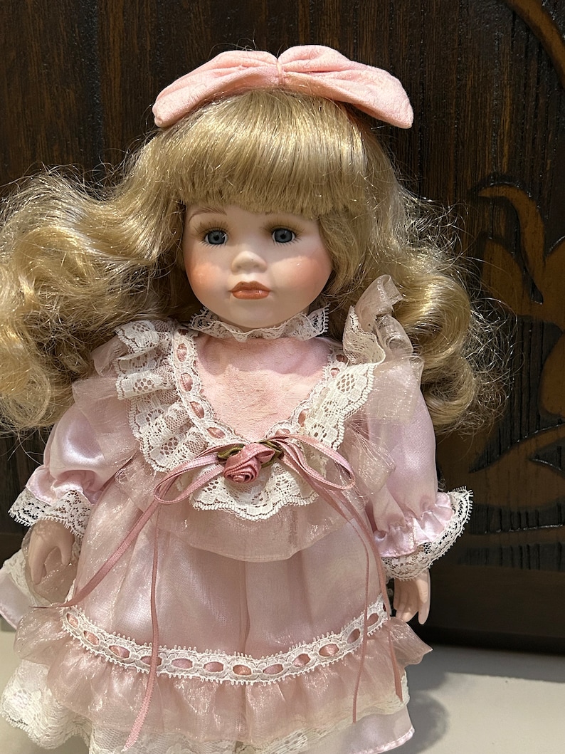 May include: A porcelain doll with blonde hair and a pink dress with lace trim. The doll is wearing a pink bow in her hair.