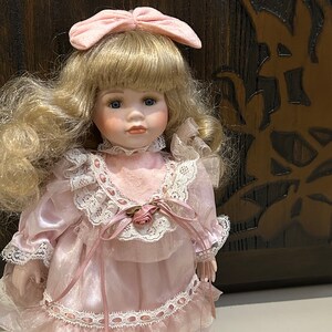 May include: A porcelain doll with blonde hair, wearing a pink dress with white lace trim and a pink bow in her hair.