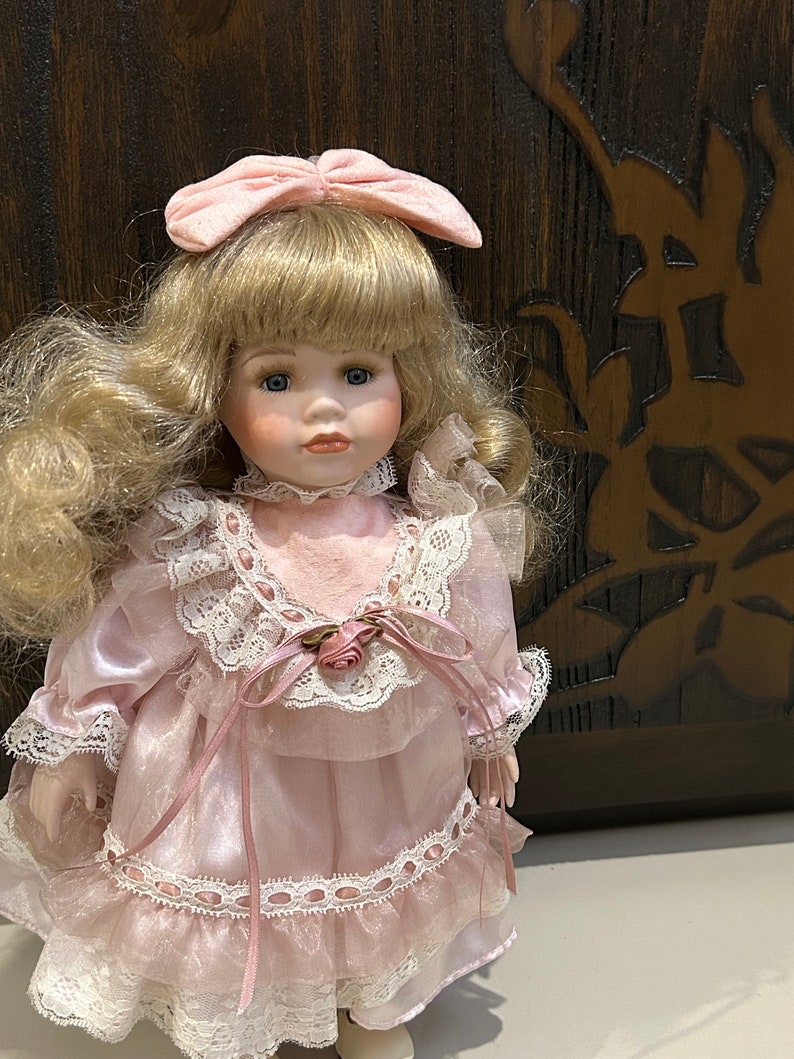 May include: A porcelain doll with blonde hair, wearing a pink dress with white lace trim and a pink bow in her hair.