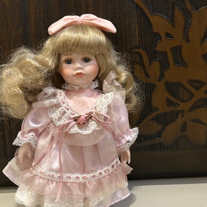 May include: A porcelain doll with blonde hair and blue eyes, wearing a pink dress with lace trim and a pink bow in her hair.