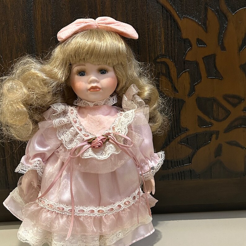 May include: A porcelain doll with blonde hair and blue eyes, wearing a pink dress with lace trim and a pink bow in her hair.