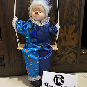 May include: A vintage clown doll in a blue satin outfit with a matching hat, seated on a wooden swing. The clown has a white ruffled collar, a red nose, and a painted face. The doll's outfit features polka dots and pom-poms.