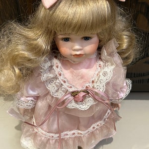 May include: A porcelain doll with blonde hair and a pink dress. The doll is wearing a pink bow and has blue eyes.
