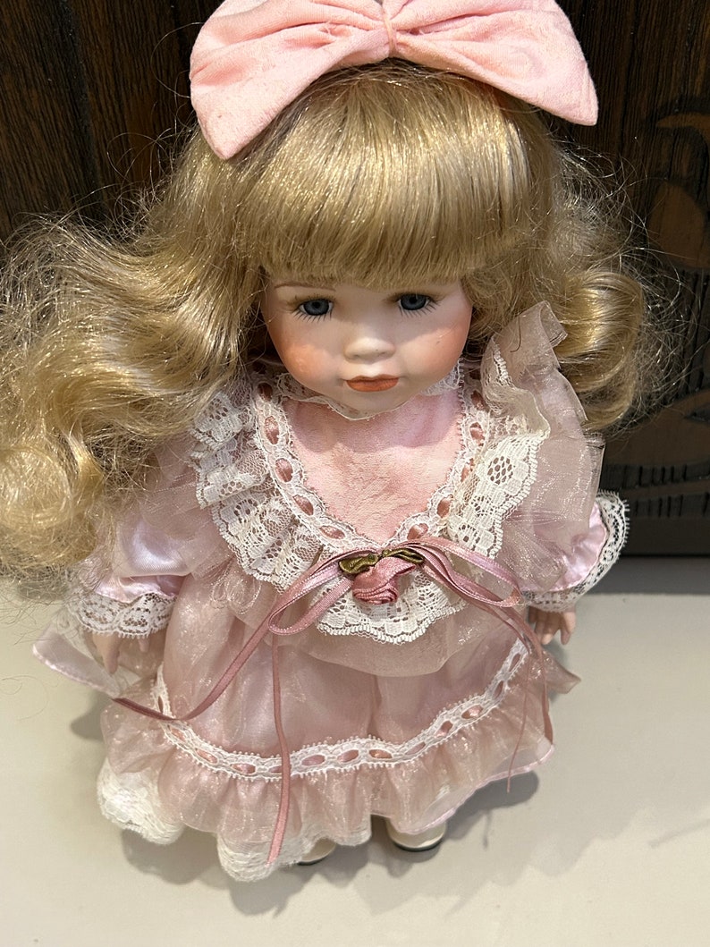 May include: A porcelain doll with blonde hair and a pink dress. The doll is wearing a pink bow and has blue eyes.