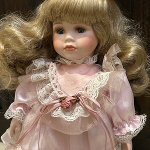 May include: A porcelain doll with blonde hair, blue eyes, and a pink dress with lace trim. The doll is wearing a pink bow in her hair.