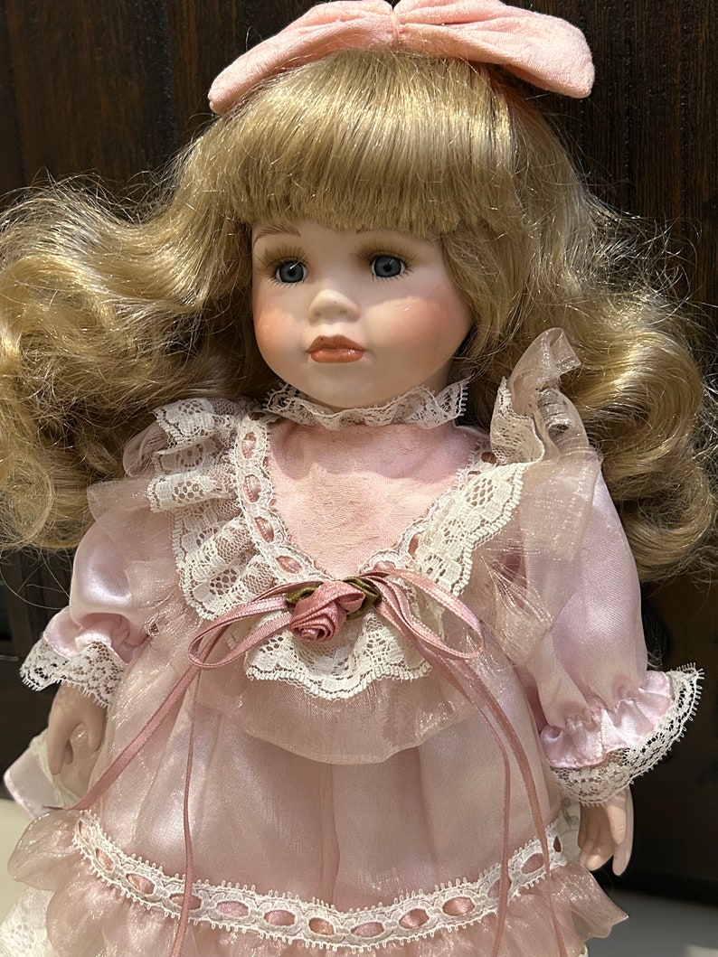 May include: A porcelain doll with blonde hair, blue eyes, and a pink dress with lace trim. The doll is wearing a pink bow in her hair.