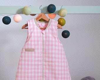 Könnte beinhalten: Ein rosa-weiß karierter Schlafsack mit Reißverschluss hängt an einem Holzbügel. Der Schlafsack ist aus weichem, atmungsaktivem Stoff gefertigt und perfekt, um Babys nachts warm und bequem zu halten.