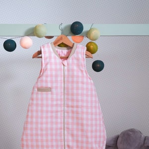 May include: A pink and white gingham sleep sack with a zip closure hangs on a wooden hanger. The sleep sack is made of soft, breathable fabric and is perfect for keeping babies warm and comfortable at night.