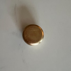 May include: A small, round, metallic gold-colored object. The top surface is slightly scratched and reflects light. The object appears to be a lid or cap, possibly for a container. The background is a white surface with subtle veining.