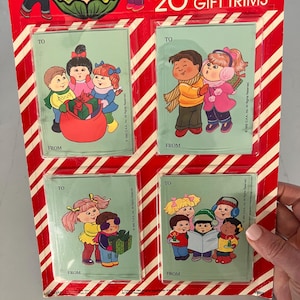 May include: A package of 20 Cabbage Patch Kids gift trims. The package has a red and white striped border. Each gift trim features a different illustration of Cabbage Patch Kids characters. The text "TO" and "FROM" are printed on each trim.
