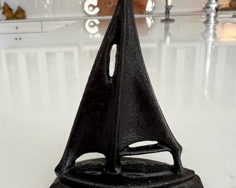May include: A dark brown, textured cast iron sailboat figurine featuring a single tall sail with a cutout opening. The boat rests on a solid, uneven base, showcasing a rugged, weathered surface finish throughout the entire metal structure.