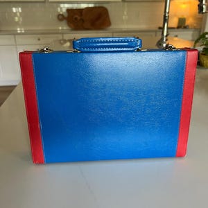 May include: A rectangular briefcase featuring a bright blue main panel with vibrant red vertical side accents. The case includes a matching blue top handle and silver-toned metal hardware attachments at the corners for the handle and closure mechanisms.