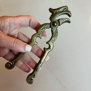 Puede incluir: Un cascanueces antiguo de latón con detalles ornamentados en forma de volutas. El cascanueces tiene un diseño de palanca con mangos curvos y una cabeza decorativa. El latón tiene una pátina ligeramente envejecida, dándole un aspecto vintage.