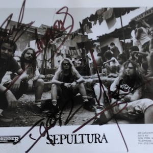 May include: Black and white photo of a band, Sepultura, signed by the band members. The band members are sitting on a concrete surface in front of a building. The photo is signed by the band members in black ink.