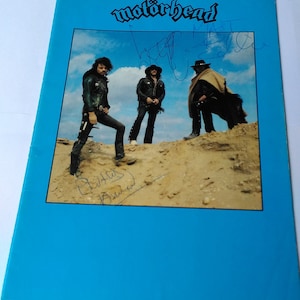 Puede incluir: Un póster azul con la banda Motörhead. Los miembros de la banda están fotografiados en un paisaje desértico, vistiendo chaquetas y sombreros de cuero negro. El nombre de la banda está escrito en una fuente estilizada en la parte superior. El póster está firmado por los miembros de la banda.