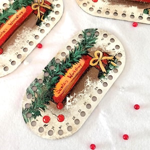 Könnte beinhalten: Ovaler Adventskalender mit dem Text "Santa's Workshop" und den Zahlen 1-31. Der Kalender ist mit einem roten und goldenen Geschenk, grünen Tannenzweigen und roten Perlenakzenten verziert. Der Hintergrund ist weiß.
