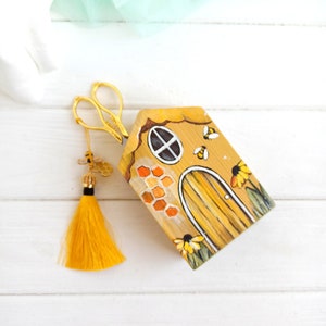 May include: A small wooden house painted to look like a beehive with a yellow door, a window, and bees and flowers painted around it. A pair of gold scissors and a yellow tassel are also in the image.