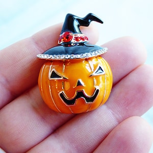 May include: A black witch hat with rhinestones sits atop an orange pumpkin with a carved jack-o'-lantern face. The pumpkin is a Halloween-themed brooch.