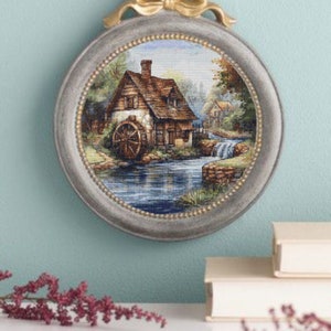 Puede incluir: Un bordado enmarcado que representa un pintoresco molino de agua. El marco redondo plateado tiene un lazo dorado en la parte superior. La obra de arte muestra un paisaje detallado con un molino de agua, un río y vegetación, creando una estética rústica y pastoral. El marco mide unos 20 cm de diámetro.