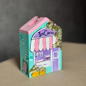 Puede incluir: Una heladería de madera pintada a mano, con un exterior turquesa y detalles en rosa y morado. La tienda tiene un toldo a rayas, un letrero que dice "Ice Cream" y una pequeña pizarra de menú. También se ven flores y plantas decorativas.