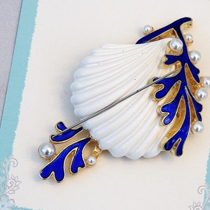 May include: A gold and blue seashell brooch with white pearl accents. The brooch features a white seashell with a blue and gold coral design.