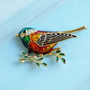 May include: A gold-tone brooch featuring a red, orange, and green bird perched on a branch with green leaves. The bird has a black eye and a single rhinestone on its head.