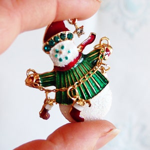 Op de afbeelding: Een groene en rode sneeuwman ornament met een gouden ketting en een witte, glinsterende afwerking.