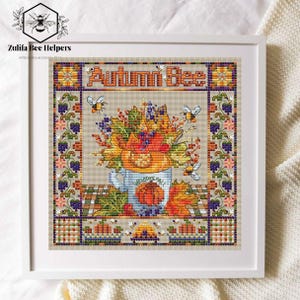 Könnte beinhalten: Gerahmte Kreuzstich-Stickerei mit den Worten "Autumn Bee". Das Design zeigt eine Tasse mit einem Kürbis, Herbstblättern und Beeren. Bienen und florale Muster umgeben das zentrale Bild. Das Kunstwerk befindet sich auf beigem Hintergrund.