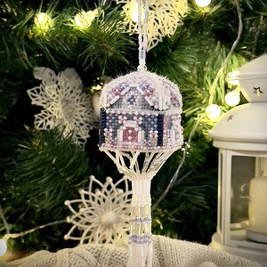 May include: A detailed, handcrafted Christmas ornament in the shape of a small house. The ornament is made with beadwork in blue, pink, and white. It hangs from a beaded string, set against a Christmas tree background.