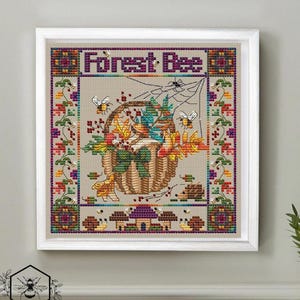 May include: Cross-stitch art in a white frame, featuring a basket filled with autumn leaves and berries. The text "Forest Bee" is at the top. Bees and a spiderweb are also depicted. The artwork has a neutral background and a decorative border.