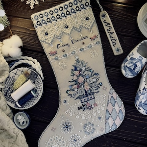 May include: Embroidered Christmas stocking with a tree design, snowflakes, and the words "Merry Christmas." The stocking is decorated with beads and a matching tag. The color scheme includes white, blue, and pink. A festive holiday decoration.