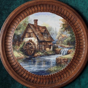 Puede incluir: Una imagen enmarcada en madera de una cabaña pintoresca con una rueda de agua y un arroyo en frente. La cabaña tiene un techo marrón y paredes blancas. La rueda de agua está hecha de madera y tiene un eje de metal. El arroyo fluye sobre rocas y tiene una pequeña cascada.