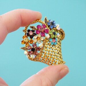 May include: A gold-tone brooch shaped like a basket overflowing with colorful rhinestones. The basket is made of a woven metal design and the rhinestones are in shades of pink, blue, green, and yellow.
